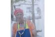 Elderly woman stands outdoors, wearing an orange dress, red headwrap, and colorful beads. A hand rests on her shoulder. Background shows trees and a white building.