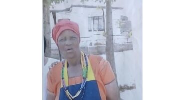 Elderly woman stands outdoors, wearing an orange dress, red headwrap, and colorful beads. A hand rests on her shoulder. Background shows trees and a white building.