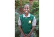 Smiling student in a green school vest and gray shirt stands in front of lush greenery, conveying a sense of happiness and pride.