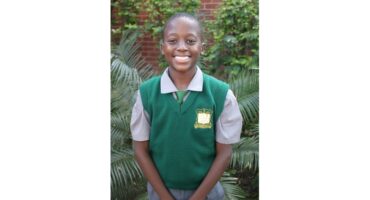 Smiling student in a green school vest and gray shirt stands in front of lush greenery, conveying a sense of happiness and pride.