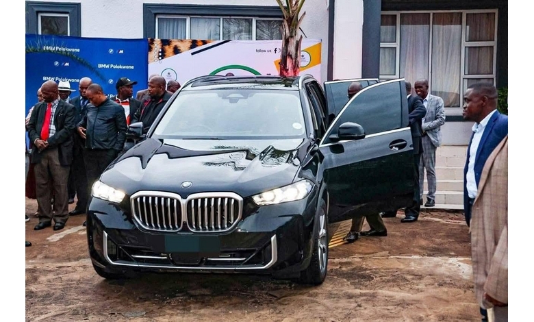 A sleek black BMW SUV is parked outdoors, surrounded by a group of formally dressed men. One man is emerging from the car, with a banner in the background.
