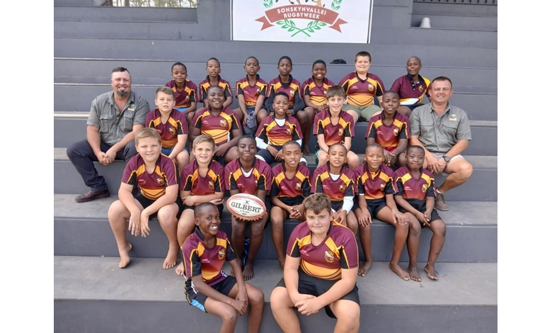 A group of young rugby players in maroon and yellow uniforms pose with two coaches. They sit on stadium steps, smiling and holding a rugby ball.