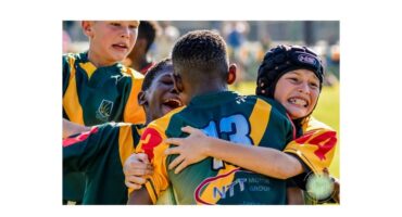Young rugby players in colorful jerseys embrace joyfully on the field, expressing excitement and camaraderie. The sunlight hints at a warm day.