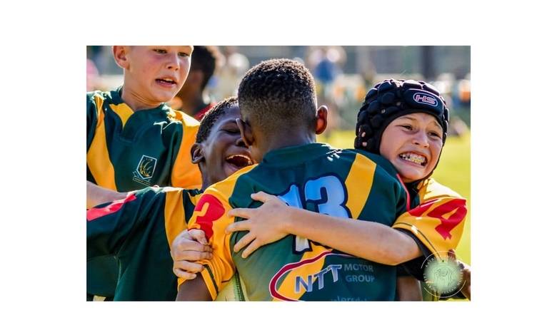 Young rugby players in colorful jerseys embrace joyfully on the field, expressing excitement and camaraderie. The sunlight hints at a warm day.