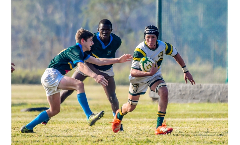 A rugby player in a green and white jersey runs with the ball, pursued by two opponents in blue and green uniforms on a grassy field, conveying intensity.
