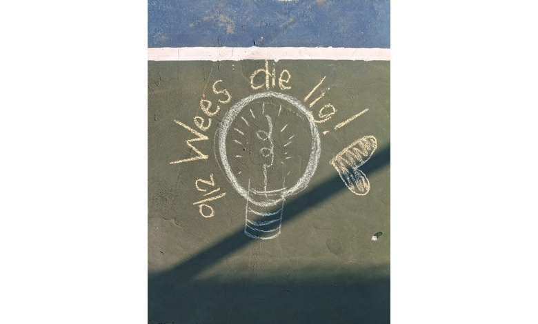 Chalk drawing on a netball court features a light bulb with the words "Wees die lig" in Dutch. Sunlight casts a shadow over part of the drawing.