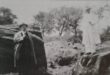 A vintage black-and-white photo shows two men in hats and coats standing on rocky terrain. One man is on a large rock; trees fill the blurry background.