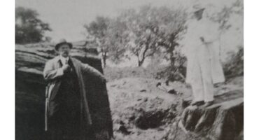 A vintage black-and-white photo shows two men in hats and coats standing on rocky terrain. One man is on a large rock; trees fill the blurry background.
