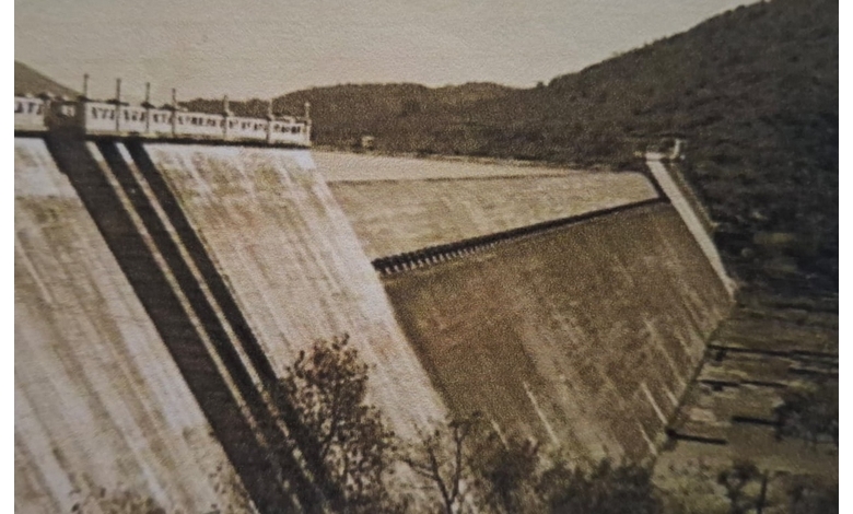 Sepia-toned image of a large, sloped concrete dam surrounded by hills and sparse vegetation. The setting appears historic, evoking a nostalgic tone.