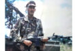 A person in camouflaged military attire stands outdoors holding a rifle. The background features trees under a blue sky, conveying a sense of readiness.
