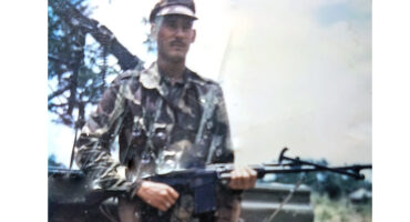 A person in camouflaged military attire stands outdoors holding a rifle. The background features trees under a blue sky, conveying a sense of readiness.