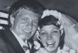 A black-and-white photo of a smiling bride and groom on their wedding day. The bride wears a floral headpiece and veil, and the groom is in a suit. Both look joyful and excited.