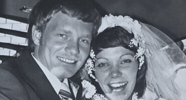 A black-and-white photo of a smiling bride and groom on their wedding day. The bride wears a floral headpiece and veil, and the groom is in a suit. Both look joyful and excited.
