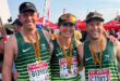 Three smiling runners in green jerseys with medals, posing arm-in-arm. Red sponsor banners and a crowd in the sunny background convey a cheerful mood.