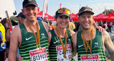 Three smiling runners in green jerseys with medals, posing arm-in-arm. Red sponsor banners and a crowd in the sunny background convey a cheerful mood.