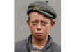 Young boy with a serious expression wearing a green beret with "ZR" insignia, gray coat. He holds a small slate. The background is plain gray.