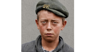 Young boy with a serious expression wearing a green beret with "ZR" insignia, gray coat. He holds a small slate. The background is plain gray.