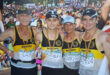 Four runners wearing "Middelburg" tank tops pose arm-in-arm, smiling proudly. They have medals around their necks, with a large, cheering race crowd behind them.