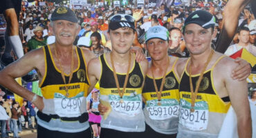Four runners wearing "Middelburg" tank tops pose arm-in-arm, smiling proudly. They have medals around their necks, with a large, cheering race crowd behind them.