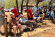 A group of men in casual clothing gather outside on a sunny day. They are sitting and standing near trees and parked cars, appearing relaxed and engaged.