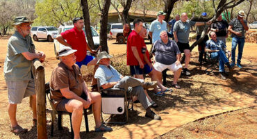 A group of men in casual clothing gather outside on a sunny day. They are sitting and standing near trees and parked cars, appearing relaxed and engaged.