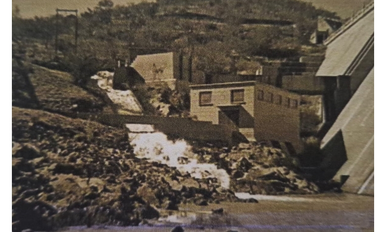 Sepia-toned image of a dam with water cascading down a rocky slope. Buildings are nestled in a hilly landscape, conveying a historical, industrial feel.