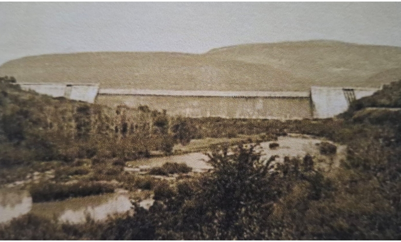 Sepia-toned photo of a large dam set against rolling hills. Lush foliage and a gently flowing river are in the foreground, evoking a tranquil atmosphere.