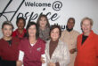A group of seven women stand smiling in front of a sign that reads "Welcome at Hospice." They wear various outfits, suggesting a warm, welcoming atmosphere.