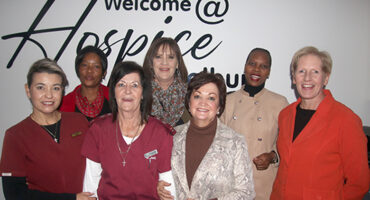 A group of seven women stand smiling in front of a sign that reads "Welcome at Hospice." They wear various outfits, suggesting a warm, welcoming atmosphere.