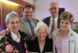 A group of five elderly people, three women and two men, smiling warmly in a room with wooden paneling. They appear to be attending a social gathering.
