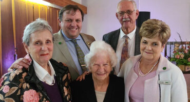 A group of five elderly people, three women and two men, smiling warmly in a room with wooden paneling. They appear to be attending a social gathering.