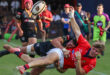 Rugby action shot: a player in a red jersey is tackled mid-air by an opponent in a black jersey. Intense focus and dynamic movement on a grassy field.