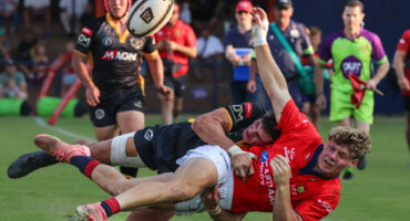 Rugby action shot: a player in a red jersey is tackled mid-air by an opponent in a black jersey. Intense focus and dynamic movement on a grassy field.