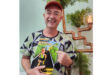A smiling man in a geometric-patterned shirt and red cap holds a colorful illustration book. Background features copper pipes and greenery, creating a cheerful vibe.