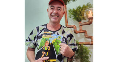 A smiling man in a geometric-patterned shirt and red cap holds a colorful illustration book. Background features copper pipes and greenery, creating a cheerful vibe.