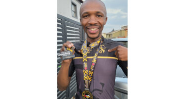 Man proudly displays medals around his neck, holding a small trophy, smiling outdoors. He's wearing cycling gear, exuding joy and achievement.