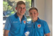 Two young men in matching blue polo shirts stand together, smiling warmly. Sunlight filters through a window behind them, creating a cheerful atmosphere.