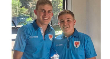 Two young men in matching blue polo shirts stand together, smiling warmly. Sunlight filters through a window behind them, creating a cheerful atmosphere.