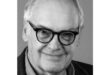 Elderly man with round glasses and light hair smiling warmly. Wears a collared shirt and jacket, set against a neutral background.