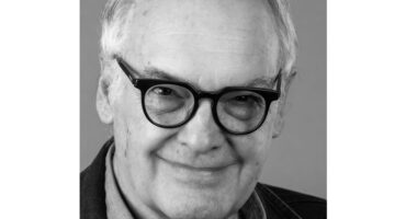 Elderly man with round glasses and light hair smiling warmly. Wears a collared shirt and jacket, set against a neutral background.