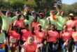 A group of smiling adults in neon athletic shirts cheerfully raise their hands outdoors. Bright and joyful atmosphere in a lush green park setting.