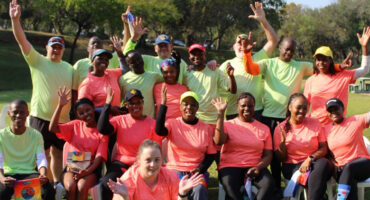 A group of smiling adults in neon athletic shirts cheerfully raise their hands outdoors. Bright and joyful atmosphere in a lush green park setting.