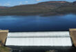 Aerial view of a large dam with water cascading over its edge. A calm reservoir lies behind, set against rugged hills and a bright blue sky.