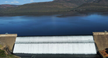 Aerial view of a large dam with water cascading over its edge. A calm reservoir lies behind, set against rugged hills and a bright blue sky.
