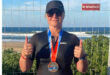 Woman in a black wetsuit and cap, wearing sunglasses, proudly shows thumbs up with a medal around her neck. She's standing by the ocean.