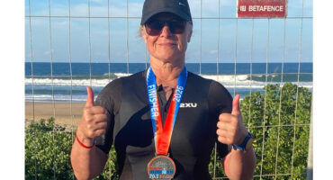 Woman in a black wetsuit and cap, wearing sunglasses, proudly shows thumbs up with a medal around her neck. She's standing by the ocean.