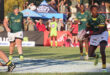 A rugby player in a green jersey prepares to pass the ball on a sunny field. Teammates and spectators watch intently, creating an energetic atmosphere.