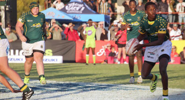 A rugby player in a green jersey prepares to pass the ball on a sunny field. Teammates and spectators watch intently, creating an energetic atmosphere.