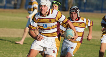 A youth rugby player in a yellow and maroon striped jersey runs with a ball, focused and determined, while teammates follow on a grassy field.