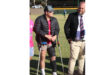 Young man stands on crutches with a leg brace, dressed casually, talking to a man in a suit on a sports field. Scene conveys support and teamwork.